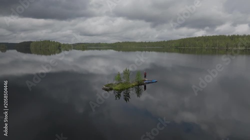 Drone footage of a pristine lake in Mujejärvi, North Karelia, Finland featuring a tiny island, a solitary kayaker, and vast coniferous forests under a moody sky.