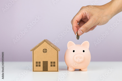 Hand putting coin in piggy bank next to small model house, conceptualizing personal savings for home purchase, property investment, and mortgage down payment