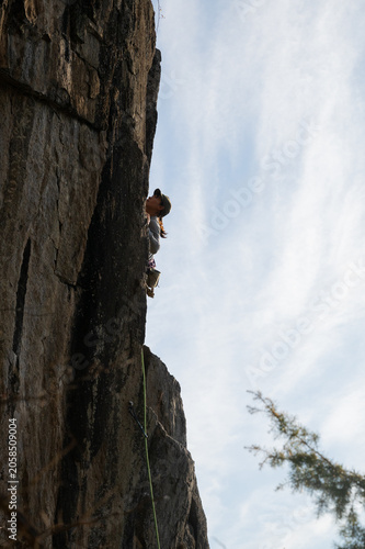 Woman rock climber scaling steep granite cliff face with green rope, low angle view against blue sky
