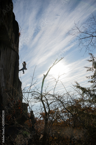 Silhouette of rock climber rappelling on steep cliff face with bare autumn trees and dramatic blue sky backlit by sun