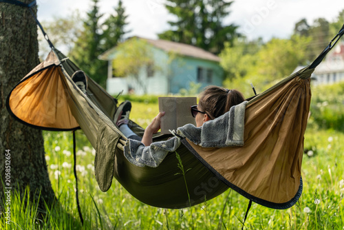 Quadro su tela A relaxed person wearing sunglasses reads a book while lying in a hammock betwee