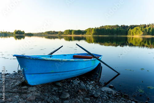 Small blue rowboat rests on a rocky lakeshore beside a still lake, with soft sunrise light, tree reflections, open sky, and a peaceful natural setting