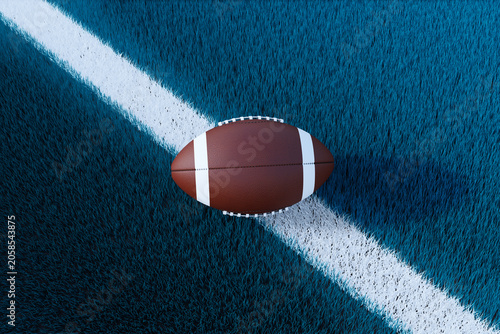 American football ball lying on clean blue turf across a white field line wit...