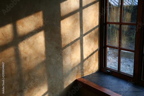 Warm sunlight streaming through a window and casting geometric shadows across a textured interior wall, creating a peaceful atmosphere and elegant natural light composition.
