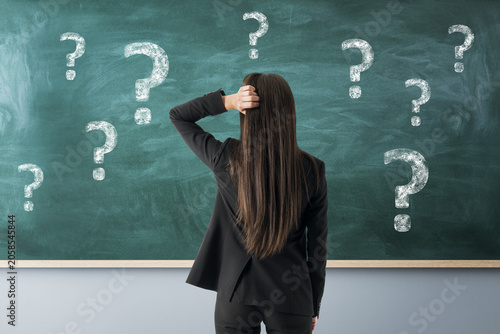 Cuadro en lienzo Confused woman standing before chalkboard filled with question marks, reflecting uncertainty and problem solving in educational setting