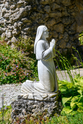 Принти на полотні White stone statue of a praying nun in a peaceful garden setting with flowers and rustic stone wall, symbolizing faith, devotion, spirituality, prayer, and serenity