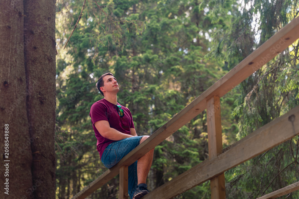 Fototapeta premium Young man resting on a wooden railing in green forest, wearing casual clothes and looking upward near a large tree with soft natural light and blurred