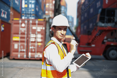 Confident logistics professional utilizes tablet for detailed inspection of busy container yard featuring numerous shipping containers and forklift during industrial operations.