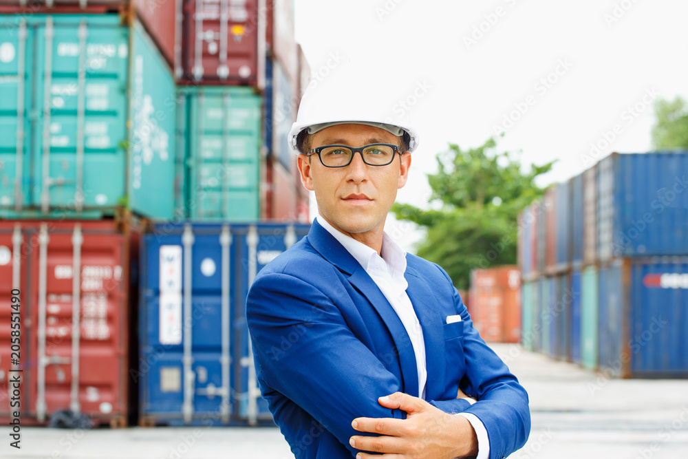 Fototapeta premium Serious logistics professional stands confidently before stacked shipping containers wearing blue suit and white hard hat, exhibiting focused demeanor during industrial operations..