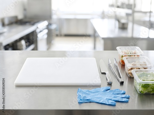 A clean commercial kitchen prep area with a separate cutting board, utensils, blue gloves, and covered food containers, showing allergen-safe food handling and contamination control.