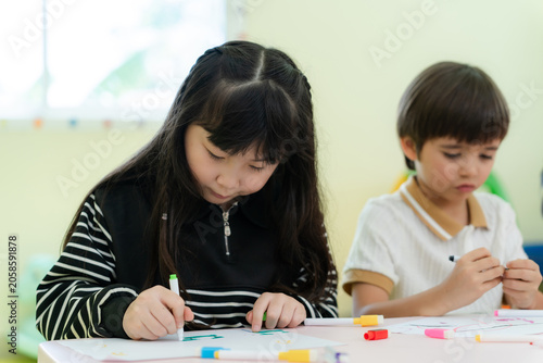 Obraz na plátně Focused little Asian girl drawing with markers next to her classmate during art