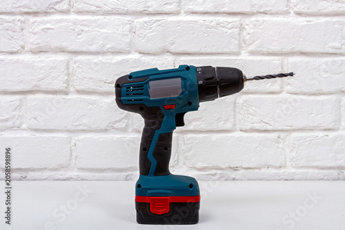 Cordless drill standing on white table against brick wall. Industrial power tool for construction or home renovation. Portable battery operated hardware for drilling and assembly