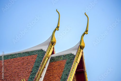 Close up of golden Chofah on traditional Thai temple roof gable with colorful tiles against blue sky background for cultural travel or Asian architectural design concept