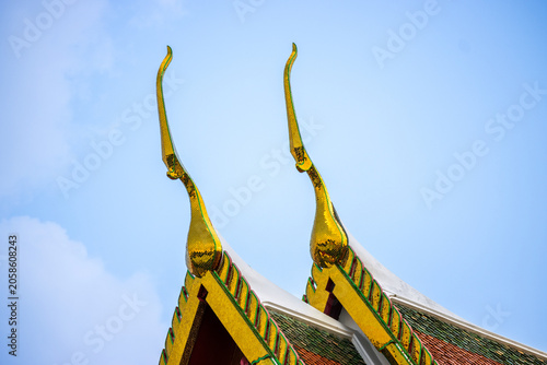 Close up of golden Chofah on traditional Thai temple roof gable with colorful tiles against blue sky background for cultural travel or Asian architectural design concept
