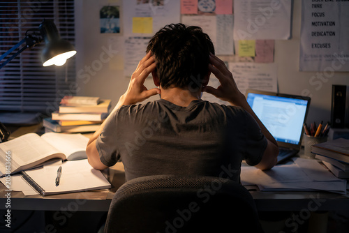 Male student studying late at night, holding his head in discomfort from a headache, books and laptop scattered across desk, realistic indoor scene.   