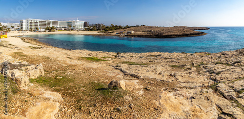Turquoise water of Vathia Gonia beach bay protected by rocky cape in winter morning. Empty sand coast with white resort hotels during sunny low season, panoramic view, Ayia Napa, Cyprus.