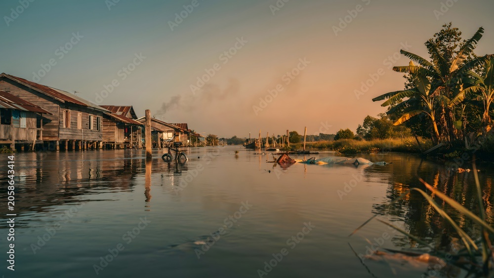Fototapeta premium Serene Village Scene with Wooden Houses and Palm Trees on Riverside at Sunset