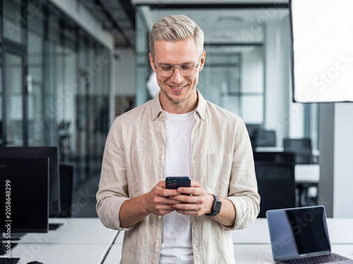 Canvastavla Smiling young businessman in glasses using a mobile phone in a bright modern office setting