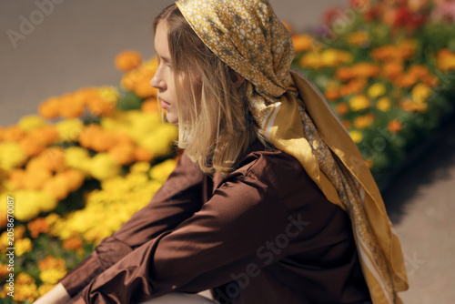 Foto Stylish blonde woman in a brown silk shirt and yellow babushka headscarf squatting by marigold flower beds at an outdoor street market