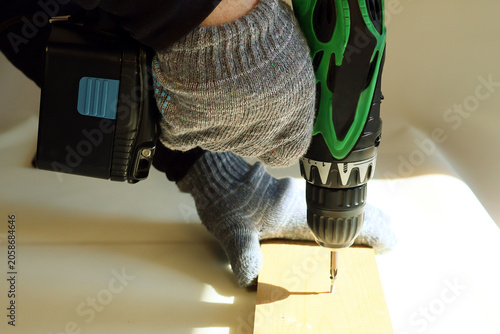 Gloved hands firmly grip green and black cordless drill. Tool drives screw into light pine wood plank. Emphasis on controlled precision and safe handling during home improvement task.