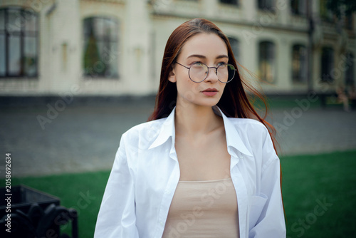 Woman with long hair wearing glasses smiles outdoors near a building on a sunny day