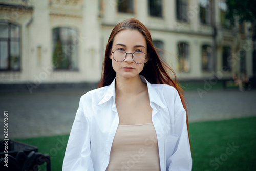 Woman with long hair wearing glasses smiles outdoors near a building on a sunny day