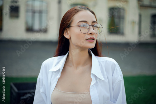 Woman with long hair wearing glasses smiles outdoors near a building on a sunny day