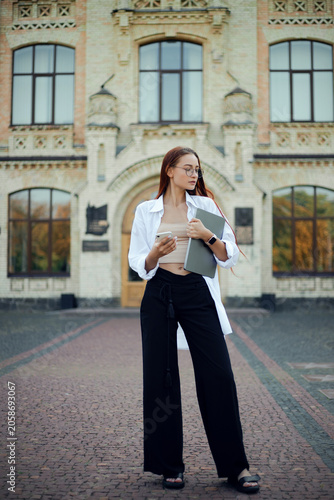 Young woman stands outside a large building holding a laptop and wearing casual clothing on a sunny day in a city