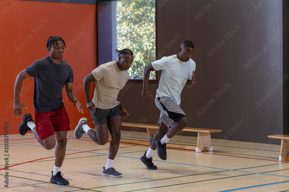 Fototapeta premium Three adult males sprinting on hardwood gym court in athletic shoes by wooden benches