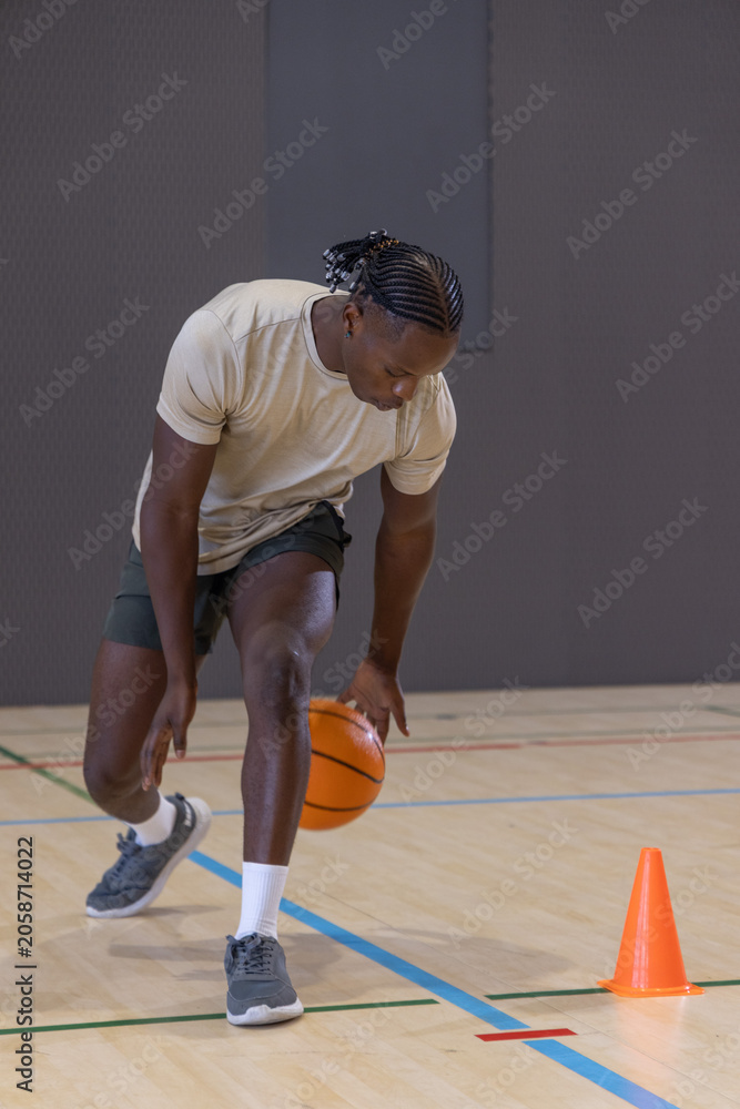Naklejka premium Basketball player is dribbling orange ball on wood court with painted lines near bright orange cone