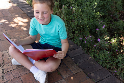 Girl child sitting cross-legged on brick path reading red book beside low hedge wearing teal tee