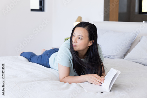 Billede på lærred Mature Asian woman lying on stomach in bedroom holding paperback book and wearin