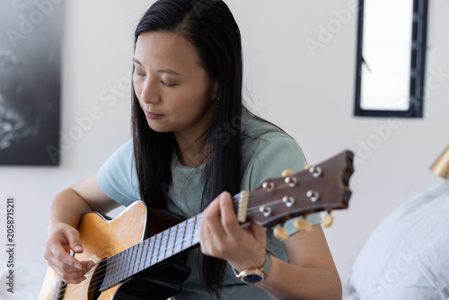 Φωτογραφία Mature Asian woman sitting on bed playing steel-string guitar in bedroom wearing
