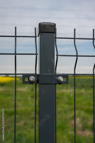 Close-Up of Modern Wire Mesh Fence Panel and Metal Post in Rural Landscape with Green Field and Yellow Rapeseed Blossoms.