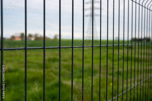 Metal security fence in focus with blurred green field and power transmission tower in background, concept of protection, boundaries, restricted access, and infrastructure.