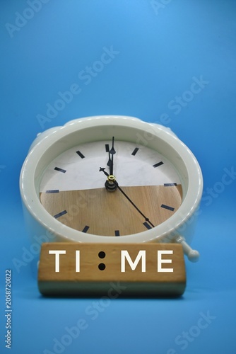 A white twin-bell alarm clock against a blue background features a wooden block in front with the word 