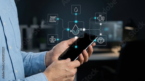 Businessman holding smartphone displaying holographic water utility management interface for monitoring industrial pipe systems and resources, digital innovation modern technology business concept