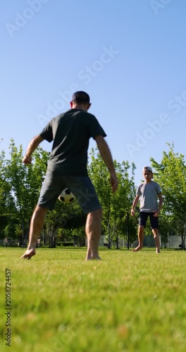 Father and son playing soccer barefoot on green grass in park. They kick ball, laugh and enjoy sunny day together. Vertical video highlights family bonding, active lifestyle and joyful childhood.