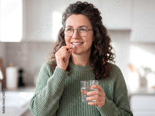 Smiling middle-aged woman with curly hair and glasses taking a pill with a glass of water in a bright kitchen.