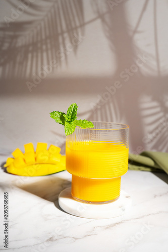 Glass of fresh mango smoothie and cut fruit on a white table with sun shadows.