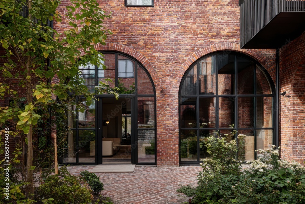 Fototapeta premium A contemporary brick building features expansive windows and is beautifully enveloped by lush greenery, all set within a vibrant urban courtyard bathed in daylight