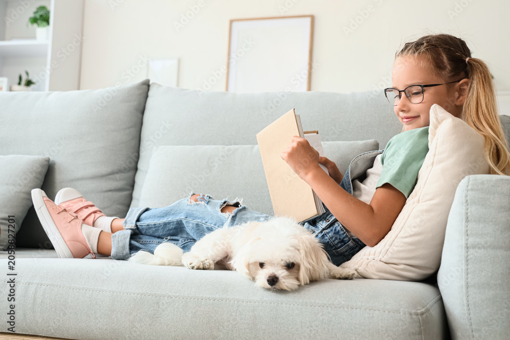 Naklejka premium Cute little happy girl with Maltese dog reading book on sofa at home