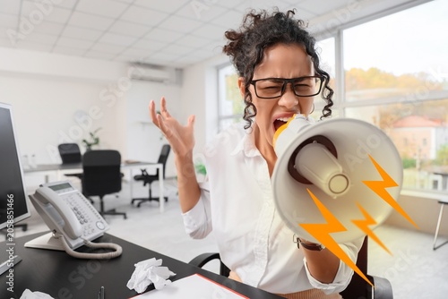 Фотография Angry businesswoman shouting into megaphone in office, expressing frustration an
