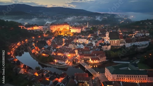 Magical evening view of Cesky Krumlov town illuminated by street lights, historic castle over Vltava river in Czechia. Cesky Krumlov at dusk with foggy hills, beautiful European heritage in Bohemia