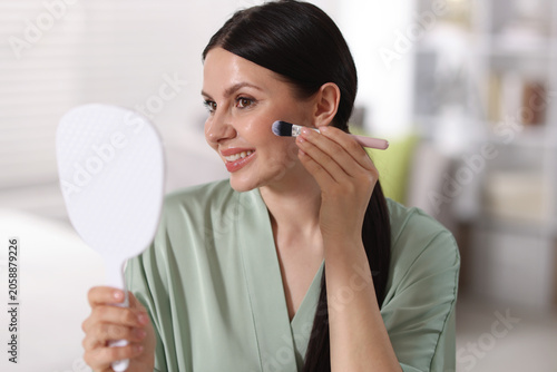 Bild auf Leinwand Young woman with mirror applying foundation with makeup brush indoors
