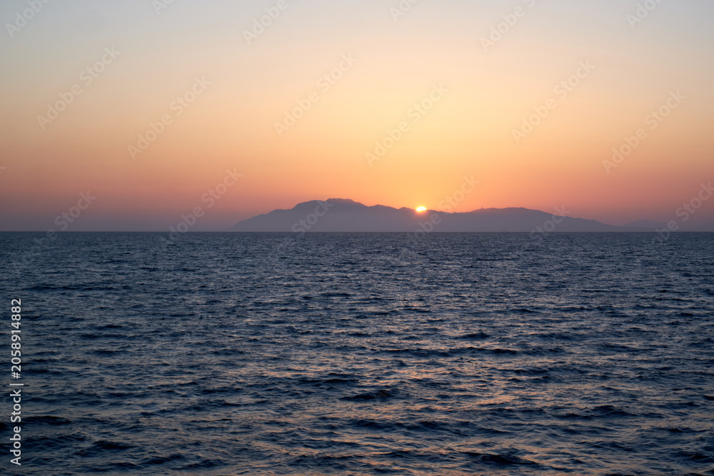 Obraz premium Peaceful serene sunset over the calm open sea with a mountainous island silhouette
