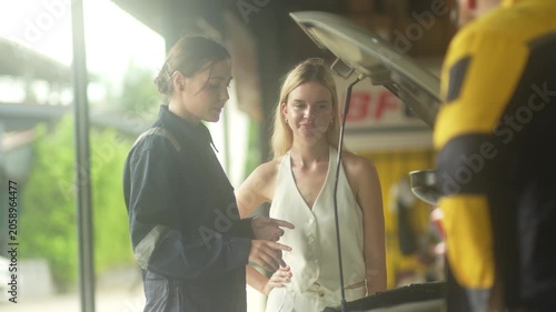 female technician in a dark uniform talks and points at the car engine, consulting a blonde female driver who smiles with confidence under an open car hood. This authentic stock imagery represents exc
