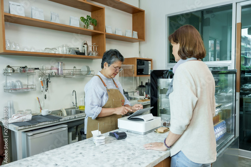 Asian senior female barista adult working at cafe counter using point-of-sale system assisting customer representing small family business with professional and friendly service