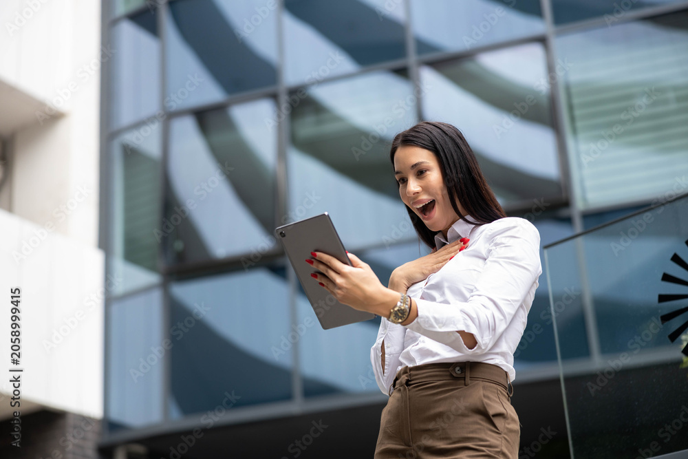 Fototapeta premium Professional woman celebrating good news, receiving success on a tablet, happy with achievements in a modern business district
