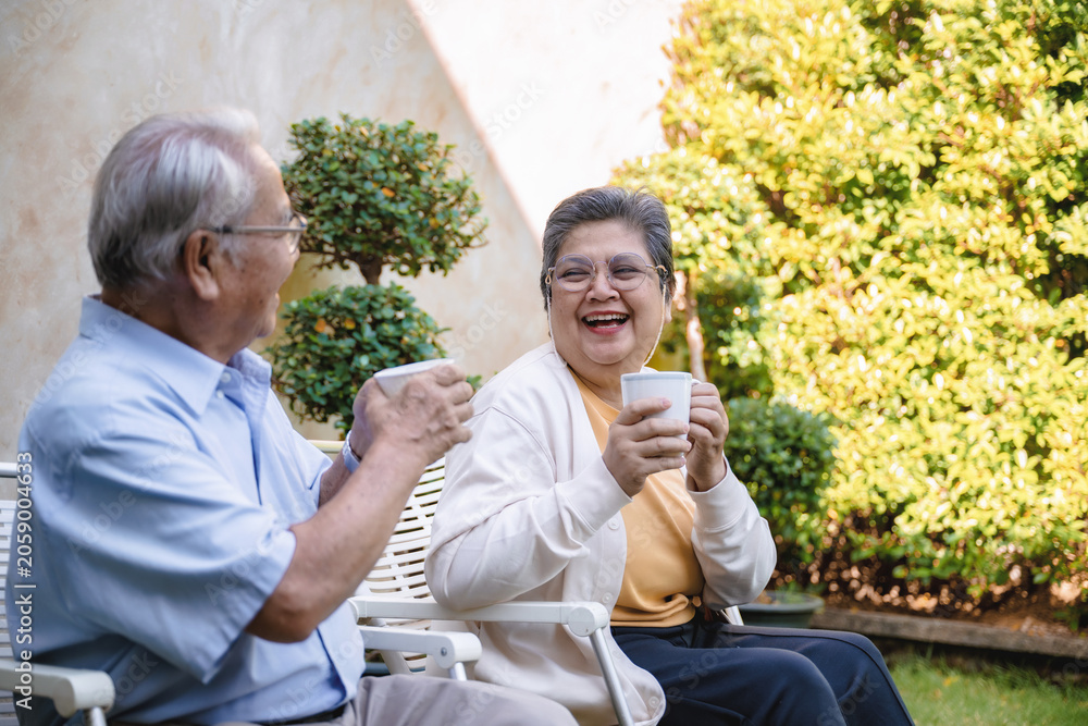 Fototapeta premium Elderly couple sits on a garden bench. Man holds a white cup, gesturing gently. Woman smiles widely, holding her cup. Sunlight illuminates green foliage in the background